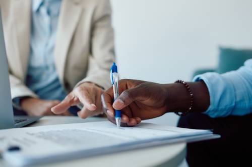 A close-up picture shows a man in a blue shirt signing an insurance contract while someone beside him explains the terms.
