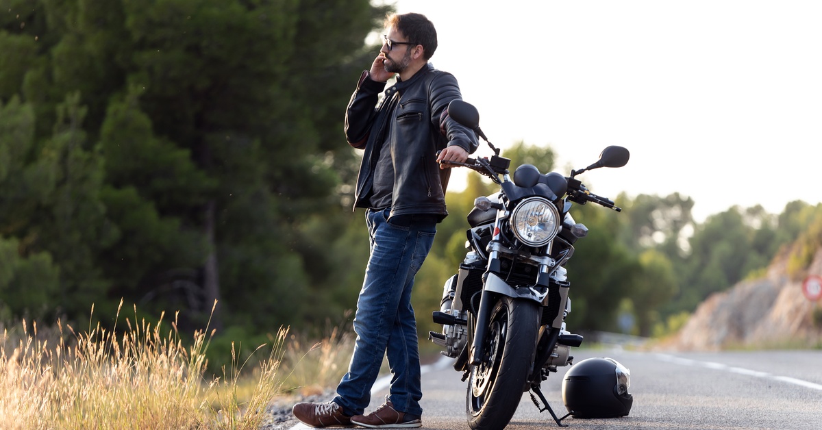 A man on the shoulder of a rural road is making a call on a cell phone and standing next to a motorcycle.