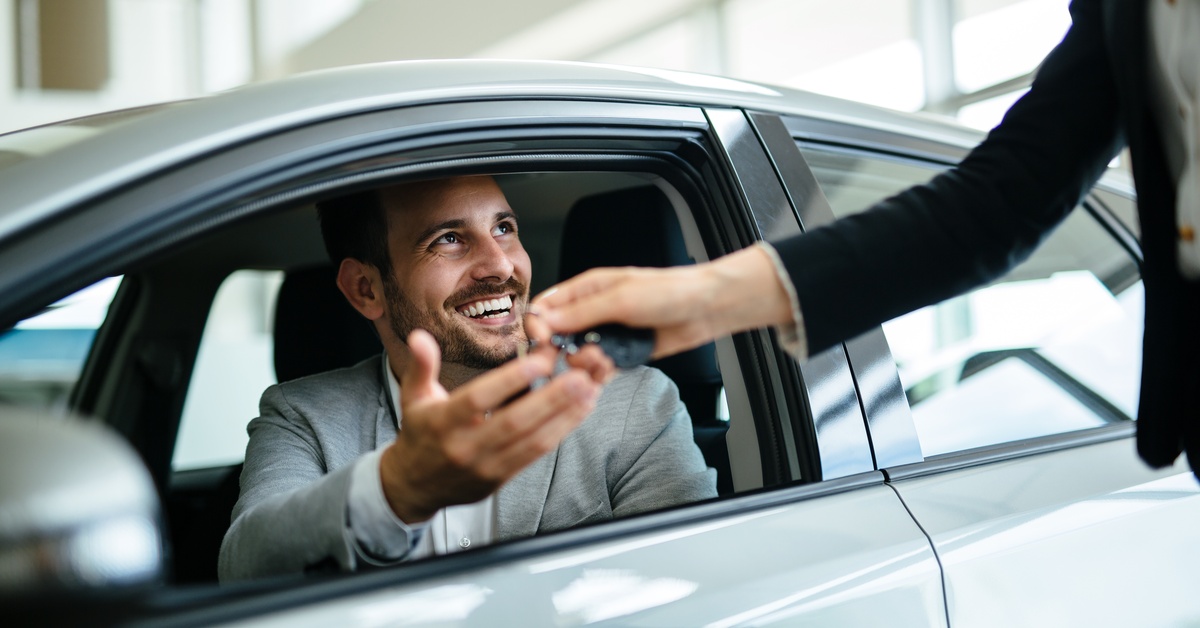Do You Need Extra Insurance Coverage for Rental Cars? A smiling man in a suit is sitting in a car, looking up at a professional handing him car keys in a warehouse.