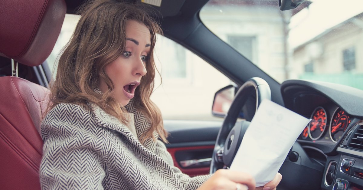 A woman sitting in a car with a red and black interior is looking at a piece of paper with a shocked expression.