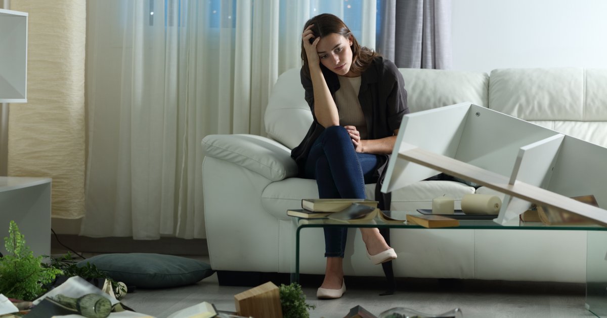 A woman in deep contemplation looks at destroyed furniture and household items strewn across a living room floor.