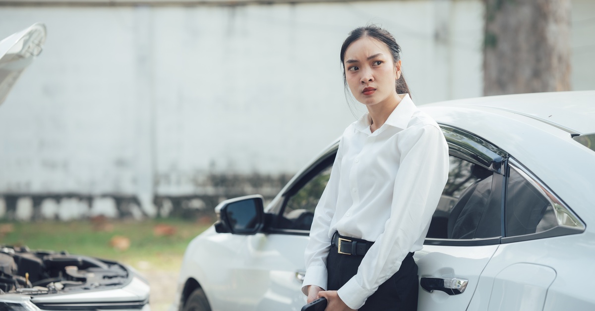 A woman in professional attire leans against a white car and makes a concerned face. Another car's hood is open in the background.
