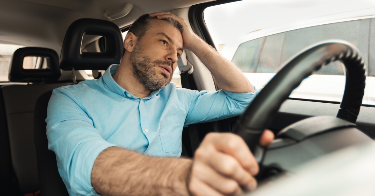 A man is sitting in his vehicle in traffic, holding one hand on the steering wheel and the other on his forehead.