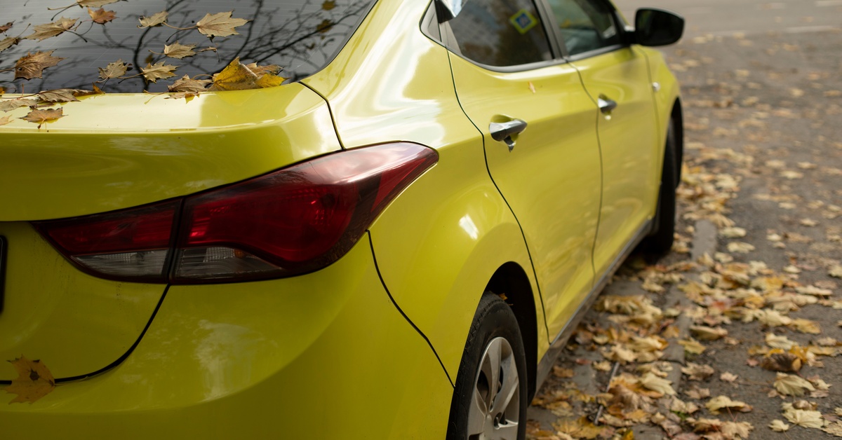  The backside of a mustard yellow car sitting on a road covered in leaves outside with grass in the distance.