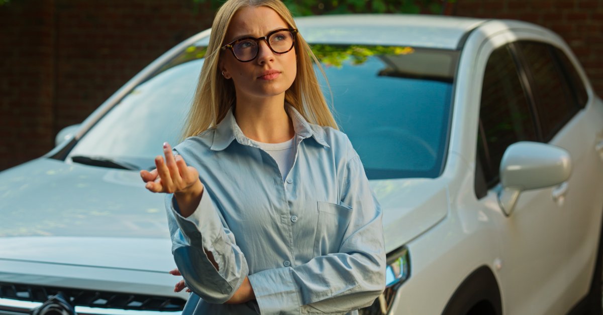 A woman in a button-up shirt and glasses is looking up with a thoughtful expression while standing in front of a vehicle.