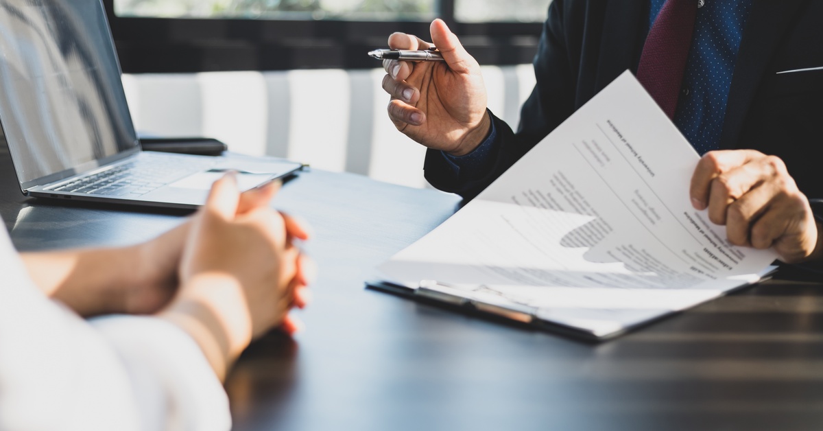 A person in a business suit is holding a pen and lifting up papers on a clipboard across from a person with their hands folded.