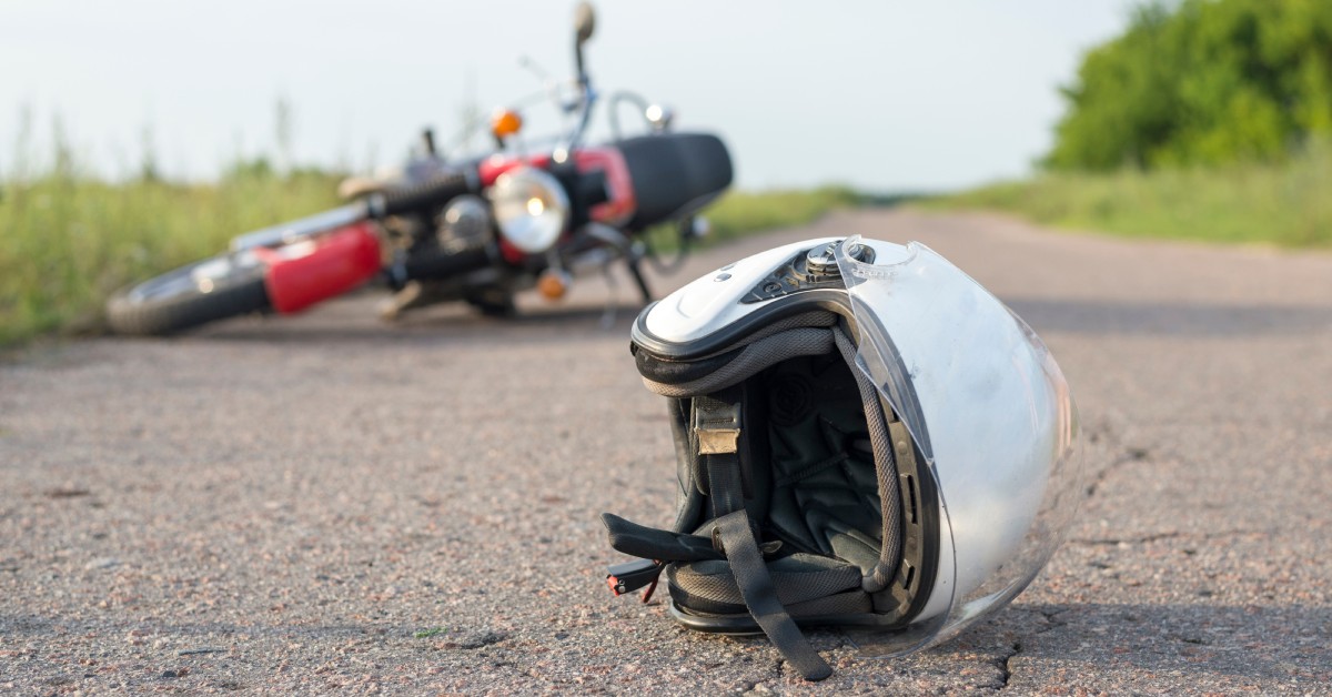 A silver helmet is lying on its side on a small road. A red-and-black motorcycle is lying on the road behind it.