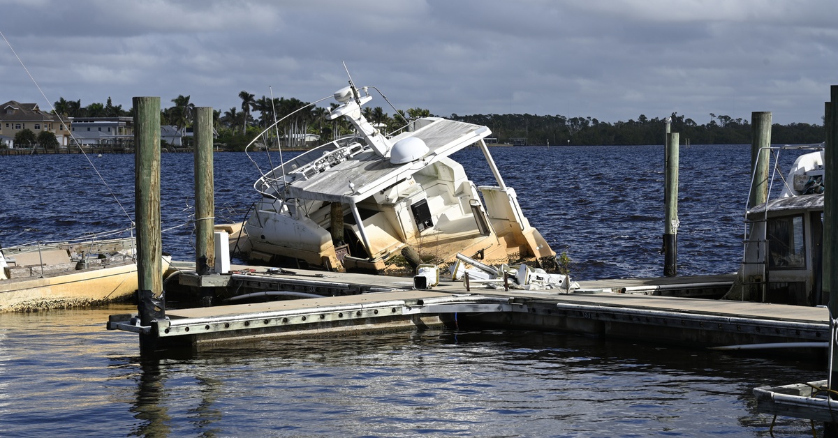 A damaged yacht sits tilted and partially sunken on the water alongside a dock with four gangways extending.