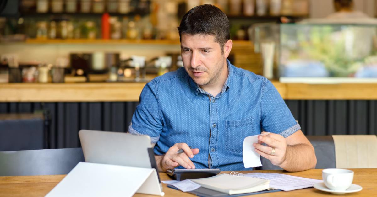 A man in a button-up shirt holds a receipt while looking at a screen. He also has a pen and is touching a calculator.