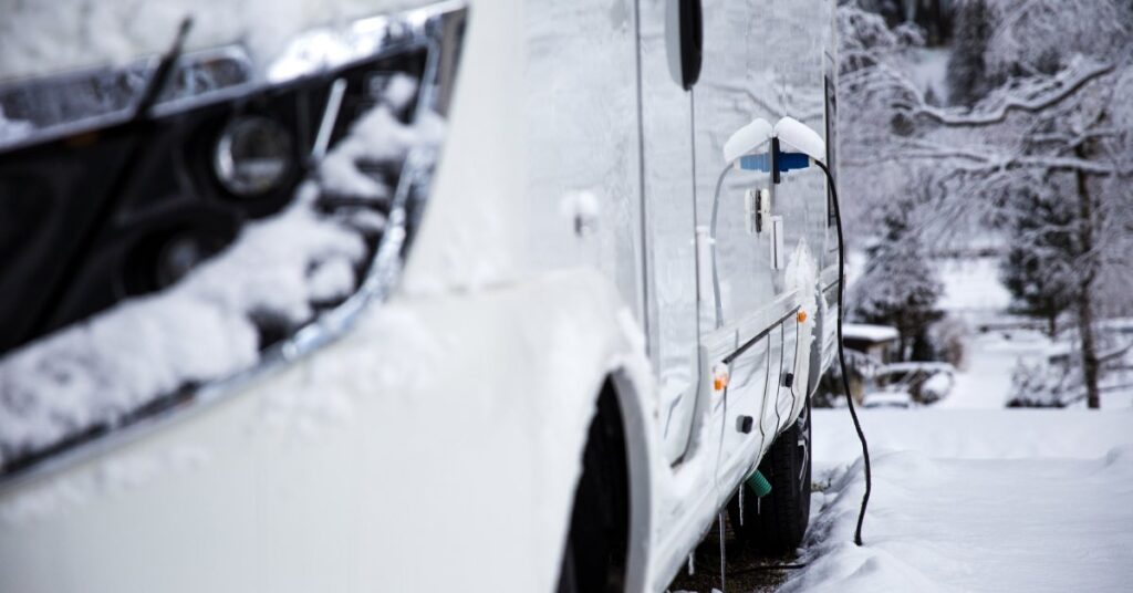 A close-up of an RV parked in the snow. Snow is built up on the headlight, and a charging cable is attached to the vehicle's side.