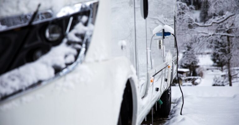 A close-up of an RV parked in the snow. Snow is built up on the headlight, and a charging cable is attached to the vehicle's side.