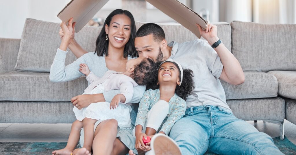 A laughing couple and their two young children sit on the floor in front of their couch holding a cardboard roof over their heads.