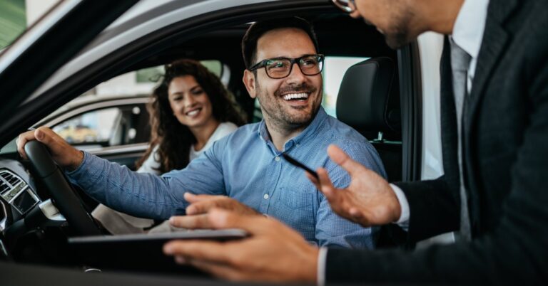 A couple smiles from inside a vehicle with the driver's-side door open. Another person leans toward them with a pen and tablet.