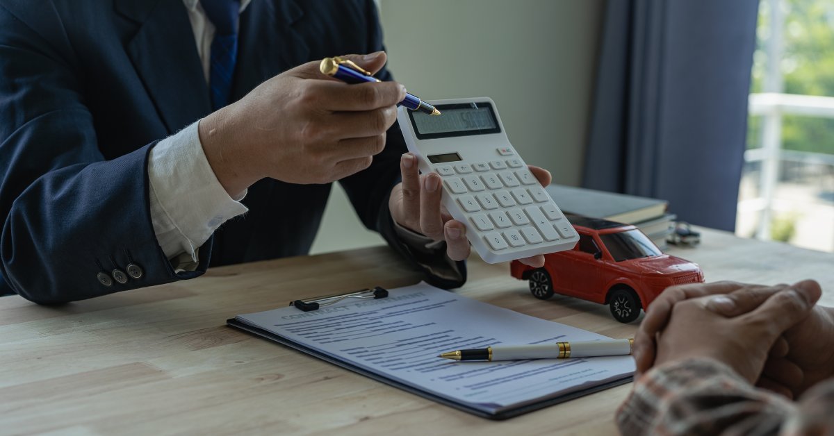 A person wearing a suit holds a calculator and points to the screen with a pen. Paperwork and a model car sit on the table.