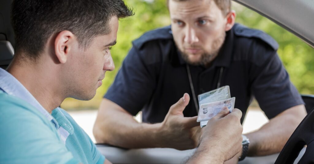 A man sitting at the wheel of a vehicle hands his insurance and registration to a police officer through the window.