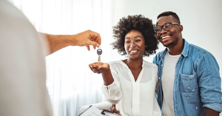 A young Black couple smiles as a person holding a clipboard hands them a set of keys in a brightly lit room with sheer curtains.