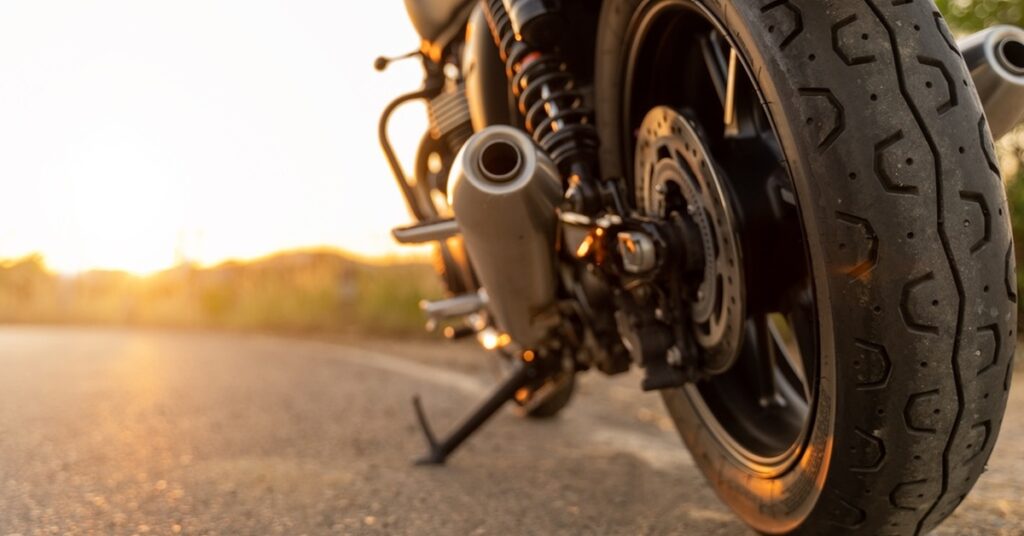 A close-up of a motorcycle parked on a road. The kickstand is down, and the sun is setting in the background.