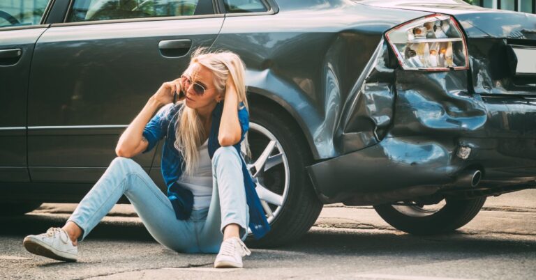 A woman sits on the ground next to a vehicle with collision damage. She is talking to someone on the phone.
