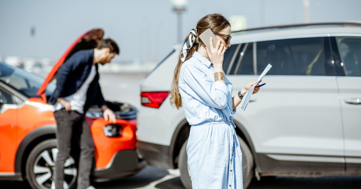A person is on the phone and looking at papers in front of a vehicle collision. Another person is leaning against a vehicle.