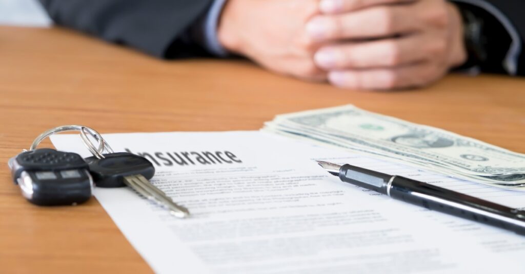 A form that reads "INSURANCE" sitting on a desk with a fountain pen, a set of car keys, and a stack of dollar bills.
