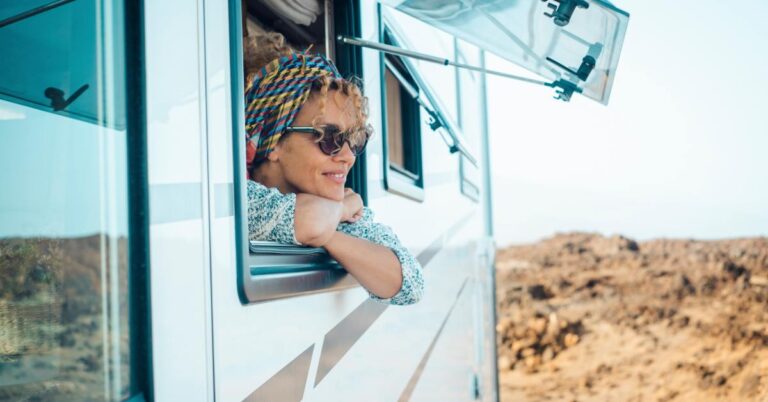 A person wearing a bandana and sunglasses leans out of the window of an RV and smiles in a desert setting.