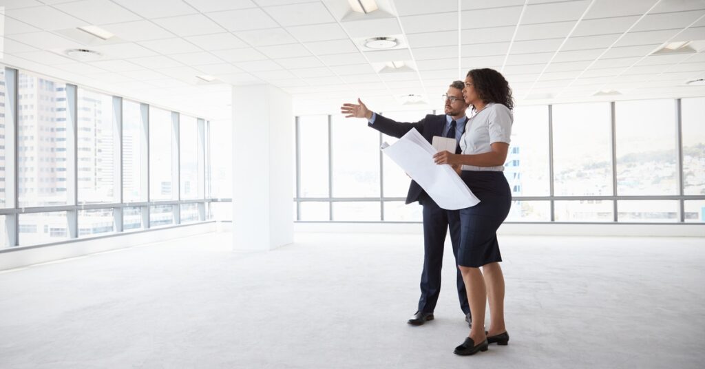 Two people stand in a vast, empty office space lined with windows. One is holding a large sheet of paper.