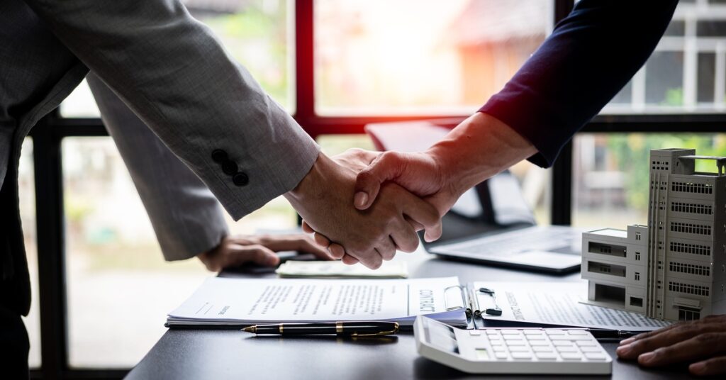 Two people wearing suits are shaking hands over a desk containing a calculator, documents, and a model of a commercial building.