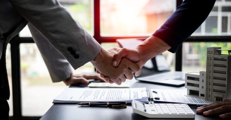 Two people wearing suits are shaking hands over a desk containing a calculator, documents, and a model of a commercial building.
