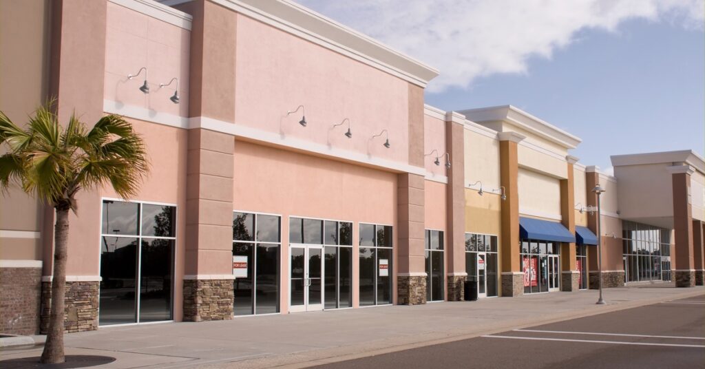 A modern strip mall with vacant storefronts with signs in the windows. A palm tree stands out front along the sidewalk.