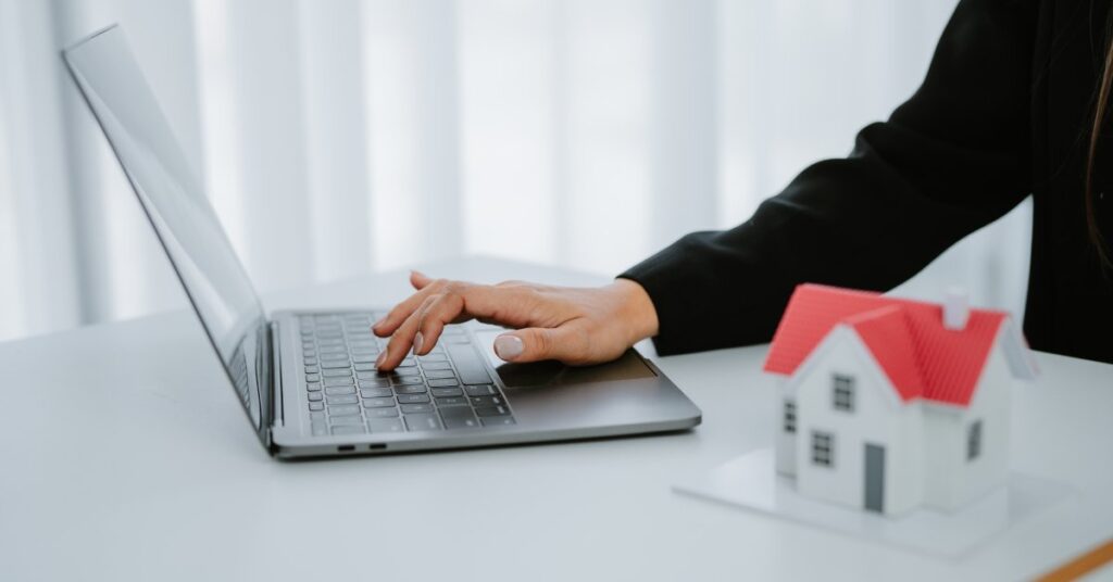 A person is pressing a key on a laptop that is sitting on a table with a miniature model of a house with a red roof.