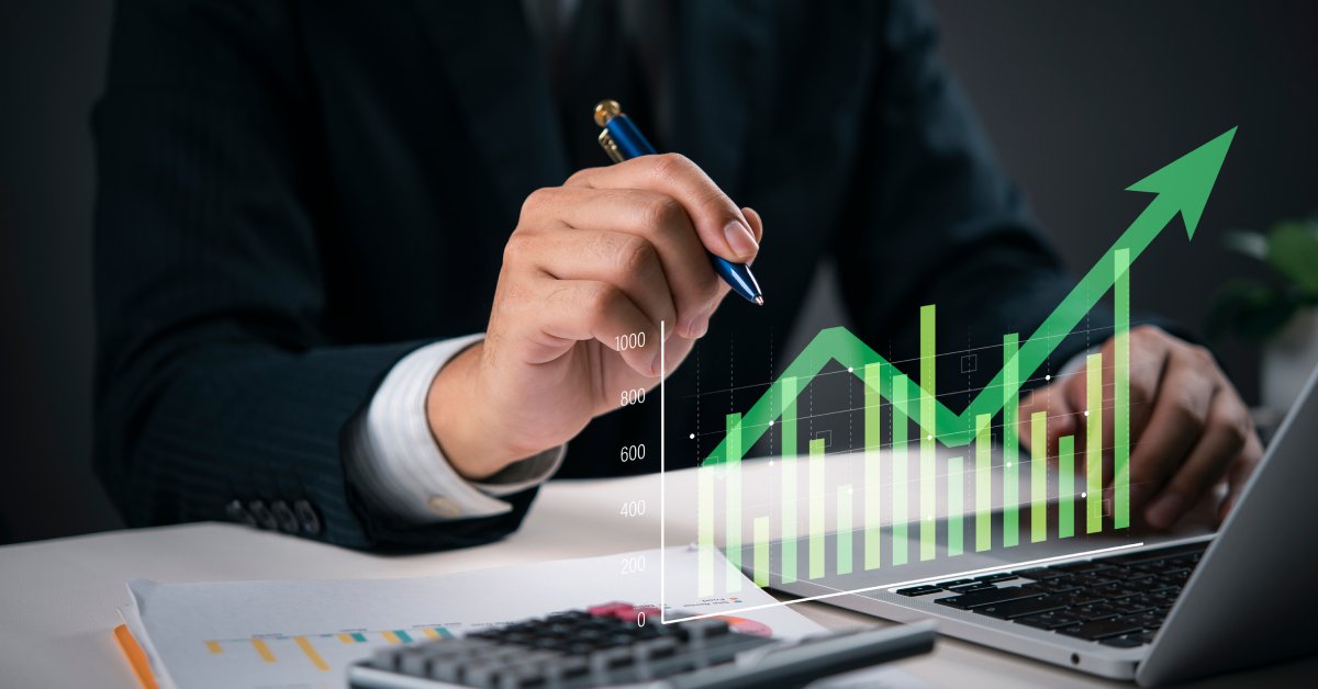 A person holds a pen in front of a calculator and a laptop. A green digital overlay shows a graph with an arrow pointing up.