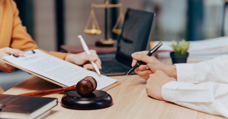 Two people sit at a desk holding pens pointed at a document on a clipboard with a gavel sitting in front of them.