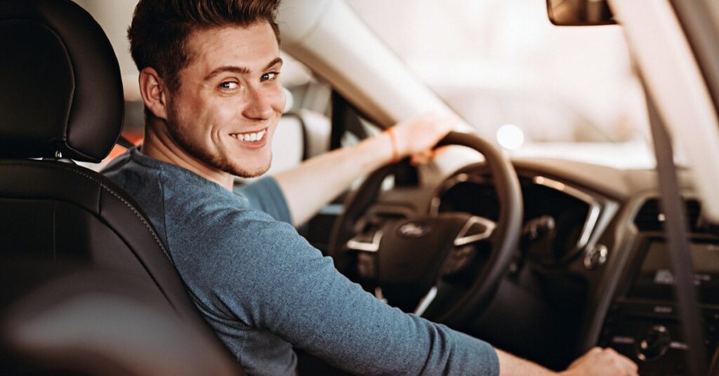 A young teen driver looks over his shoulder with one hand on the steering wheel and one on the gear shift.