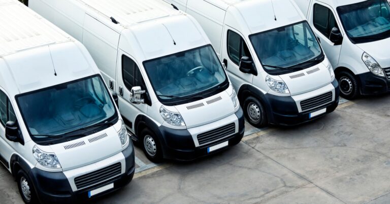 Several white delivery vans are lined up next to each other on a concrete lot with painted parking spots.