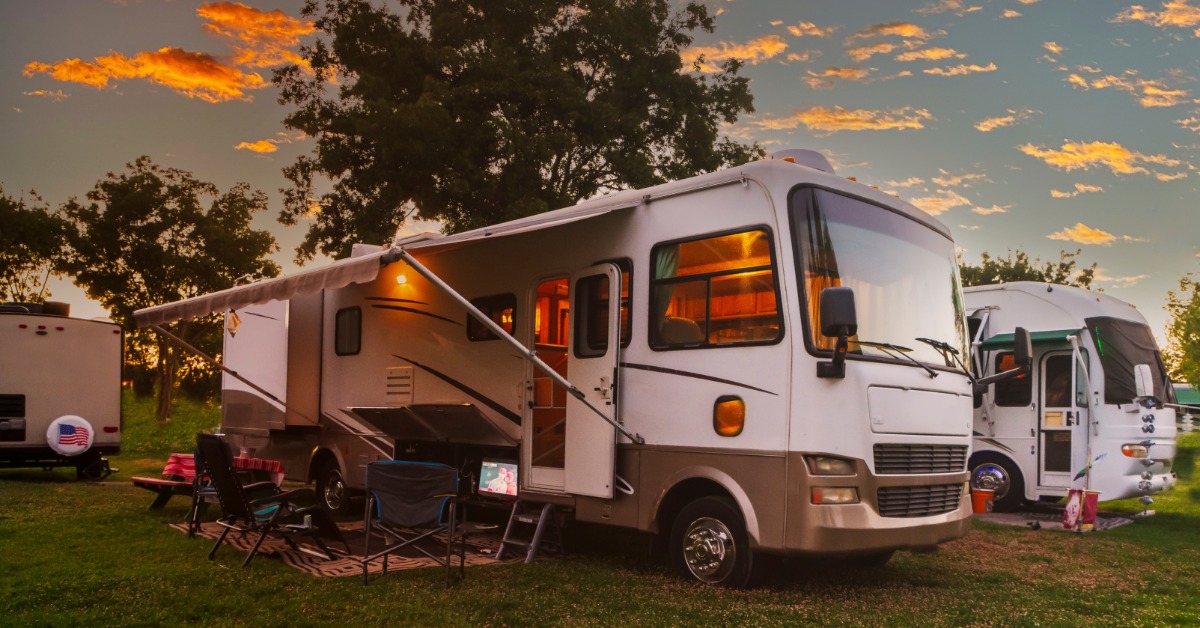Two motorhomes sit at a grassy campsite at sunset. One has its awning open with chairs and a TV set up beneath it.