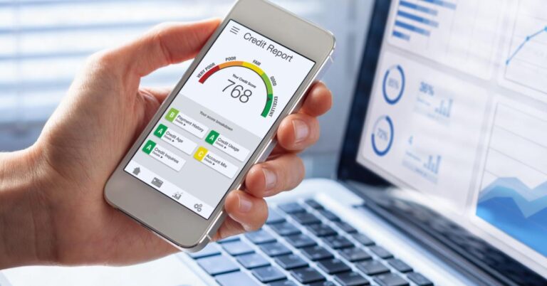 A person holds a smartphone displaying a credit report score of 768 with financial charts open on a laptop in the background.