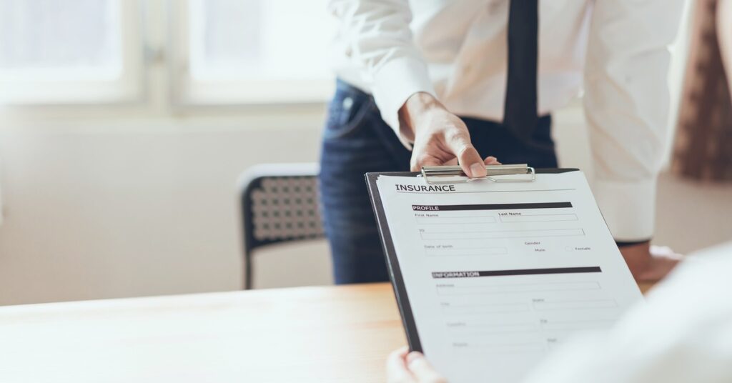A person in a white shirt and tie hands an insurance form on a clipboard to a seated client across a desk.