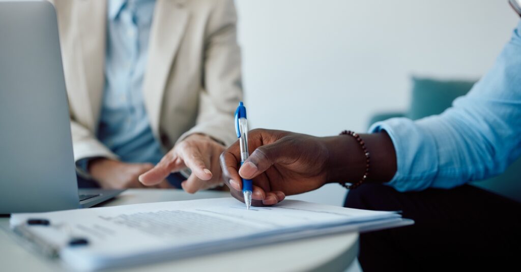 A person signs a stack of paperwork with a pen while another sits in front of a laptop during a meeting.
