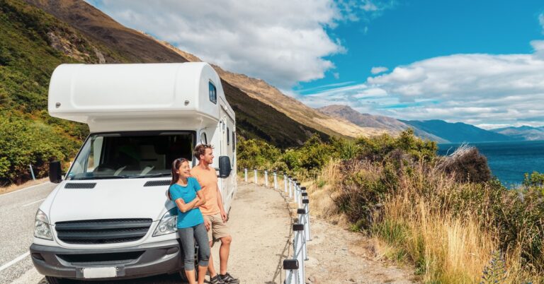 A young man and woman couple standing next to a white RV parked on the side of a road overlooking a cliff.