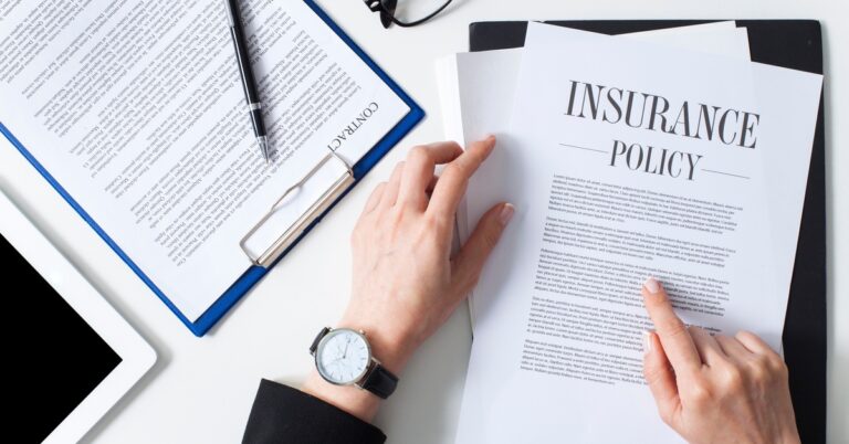 An individual in a black jacket reviewing insurance policy paperwork on a desk with glasses, a pen, and a clipboard.