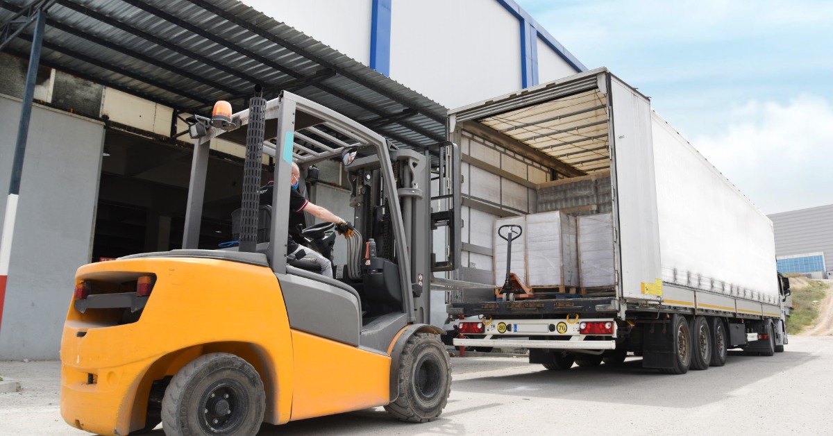 A forklift loading cargo into a semi-trailer at a warehouse dock, with pallets inside and a worker operating the vehicle.
