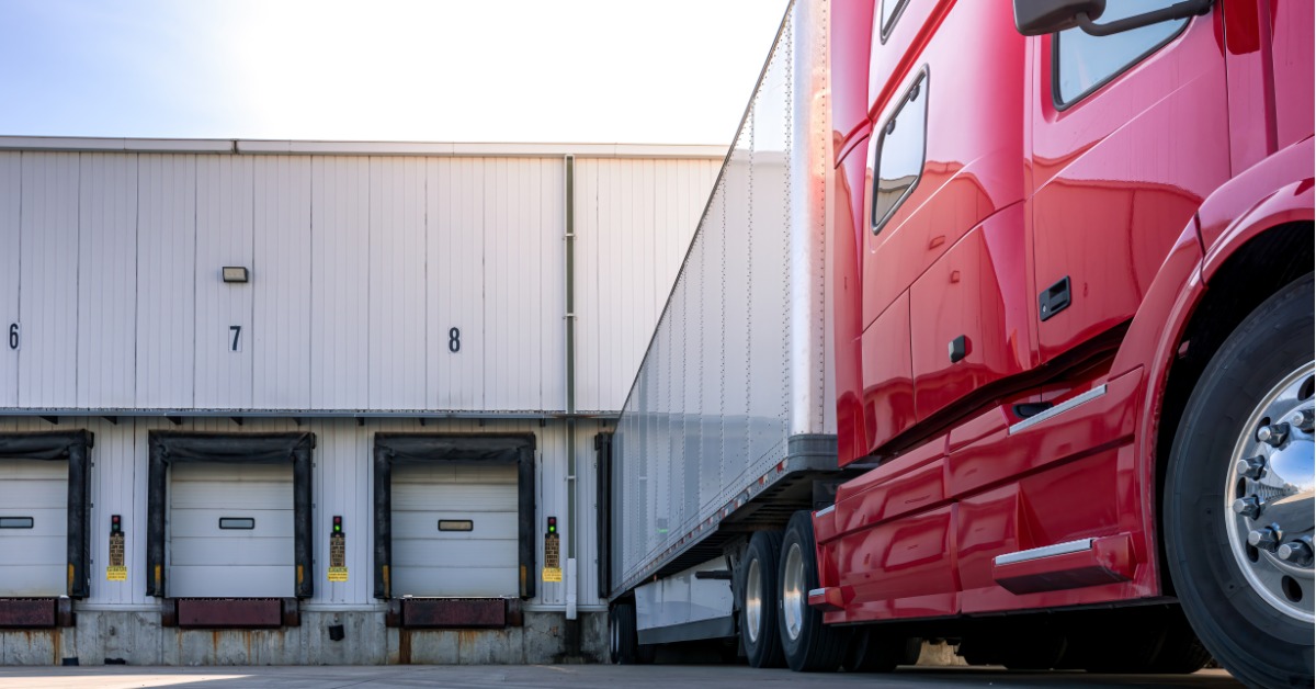 A red semi-truck with a trailer backed into a warehouse loading dock, with numbered bays and closed doors.