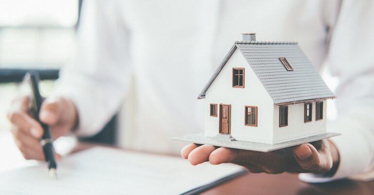 A person in a white shirt holding a small model house while using a black pen to sign paperwork on a clipboard.
