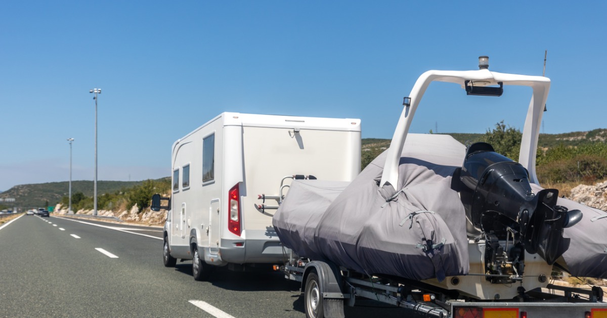 A white RV towing a covered boat trailer along an open highway beneath clear blue skies on a sunny day.