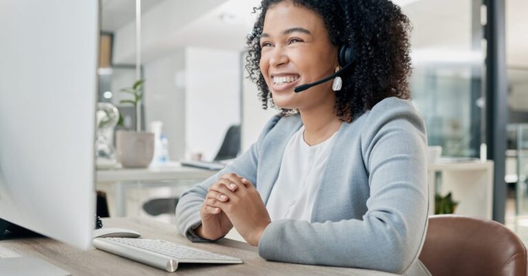 A smiling female customer service representative wearing a headset while sitting at a desk in a bright modern office.