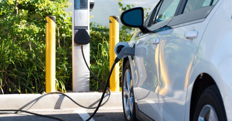 A white electric car charging at a public station beside yellow posts and green shrubs in a sunny parking lot.