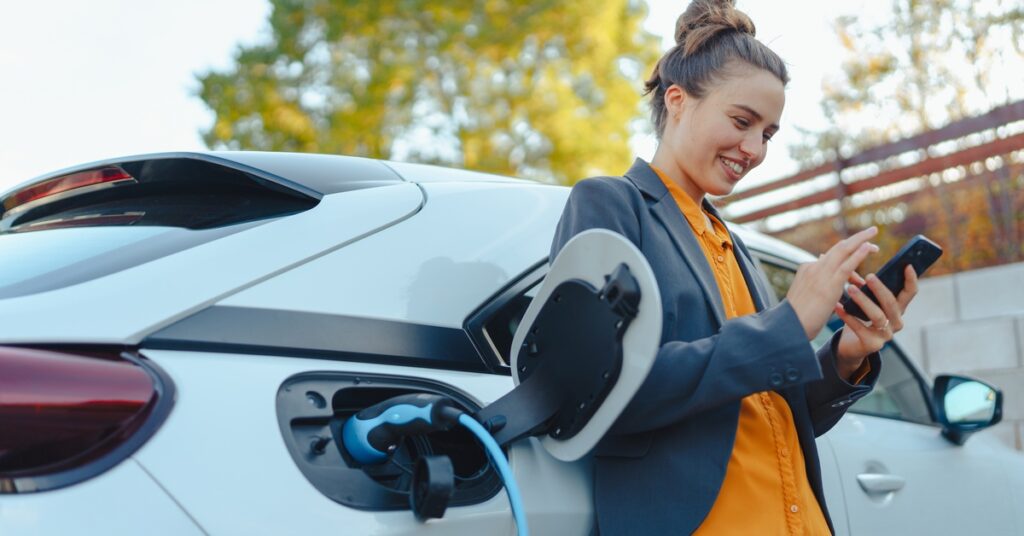 A woman using a smartphone while charging a white electric vehicle outdoors near trees on a sunny afternoon.