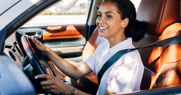 A smiling woman wearing a seatbelt drives a car with tan leather seats, viewed through the open driver's window.
