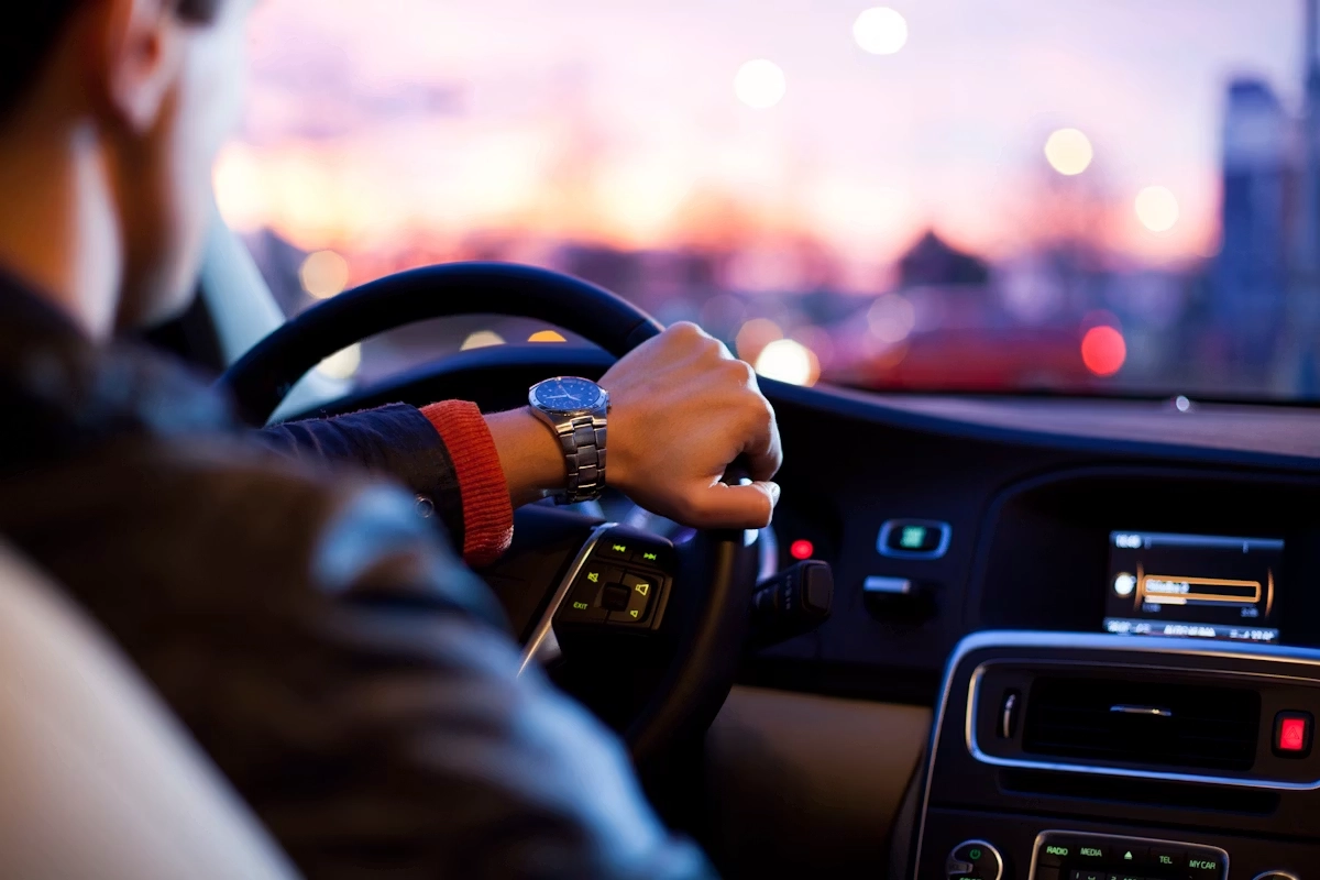 Close-up of a driver's hand on the steering wheel while driving at dusk, with city lights blurred in the background